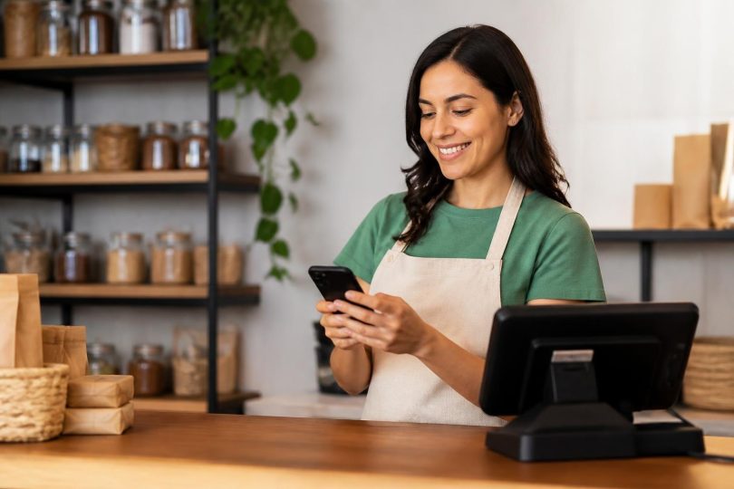 Empresário usando celular para automatizar cobranças Pix ao lado de caixa registradora
